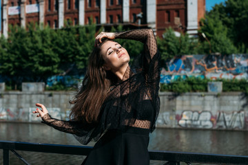Fototapeta premium beautiful girl, brunette, in the summer by the river in the city, in a black dress. standing alone by the river, on the bridge, the sun is shining in the face, closed her eyes, rest and tourism.
