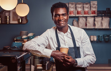 African barista smiling at camera relaxing after workday with coffee while leaning on the counter...