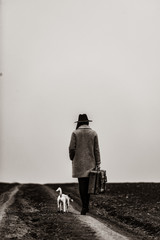 portrait of a young beautiful woman with suitcase and dog standing in the field. Image in black and white color style