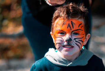 A small young boy who has his face painted to look like a tiger looking at the camera with a smile in the middle of Autumn