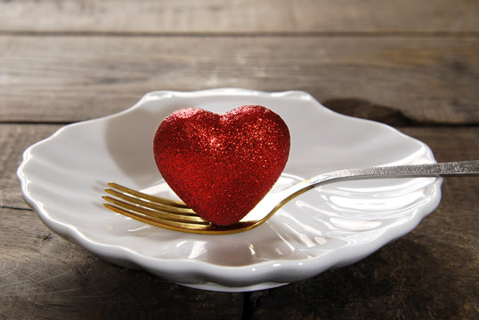 Red heart dotted with sequins and a fork on a white plate in the shape of seashells on a dark wooden background.