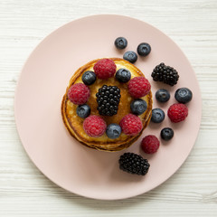 Pancakes with berries on a pink plate on a white wooden table, top view. Close-up.