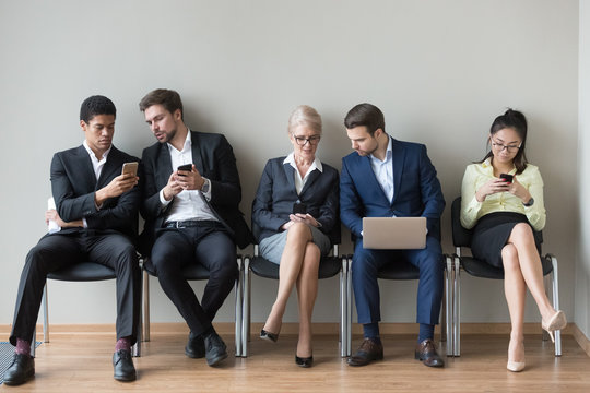 Multiethnic work applicants busy using laptops and smartphones preparing for recruiting talk, diverse job candidates sit in queue waiting for interview talking, browsing gadgets. Hiring, HR concept