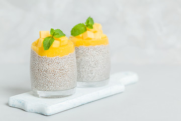Chia seeds pudding with mango puree in beautiful glasses with green mint leaves and cut fresh ripe tropical fruit.