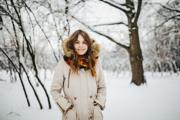 Theme is a weekend holiday in winter. Beautiful young Caucasian woman standing in snow park in jacket with hood and fur in jeans and smiling