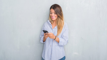 Young adult woman over grey grunge wall looking at smartphone texting a message with a happy face standing and smiling with a confident smile showing teeth