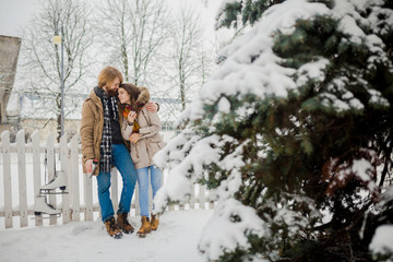 Fototapeta na wymiar Young loving couple caucasian man with blond long hair and beard, beautiful woman have fun drinking a hot drink from thermos, eating green apple in snow park near white fence and coniferous tree