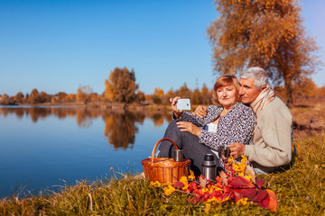 Senior couple taking selfie while having picnic by autumn lake. Happy man and woman enjoying nature...