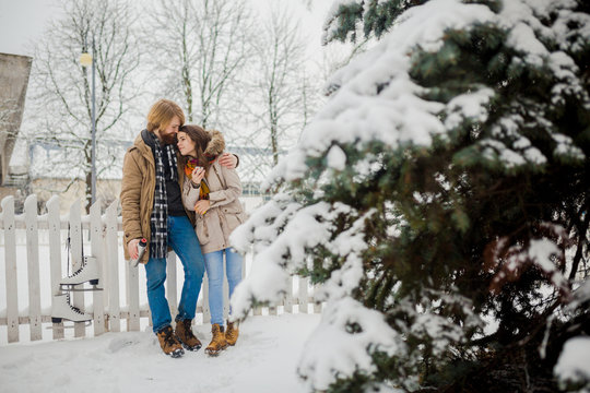 Young loving couple caucasian man with blond long hair and beard, beautiful woman have fun drinking a hot drink from thermos, eating green apple in snow park near white fence and coniferous tree