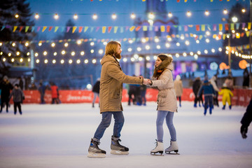 Young couple in love Caucasian man with blond hair with long hair and beard and beautiful woman have fun, active date skating on ice scene in town square in winter on Christmas Eve