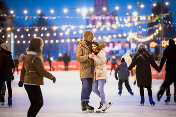 Young couple in love Caucasian man with blond hair with long hair and beard and beautiful woman have fun, active date skating on ice scene in town square in winter on Christmas Eve