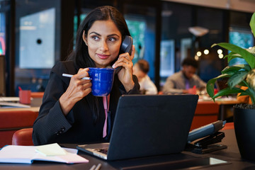 Businesswoman drinking coffee while talking on telephone in office