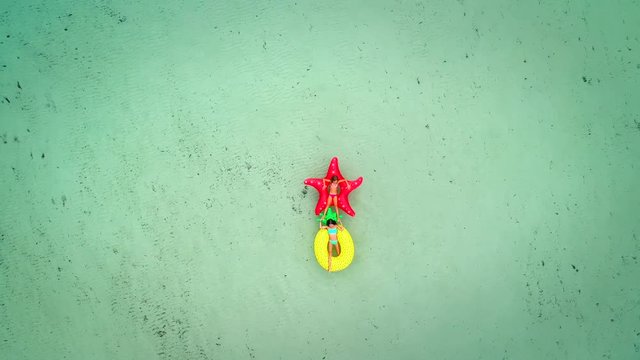 Aerial view of two girls floating on inflatable star fish and pineapple mattress on transparent sea, touching feet.