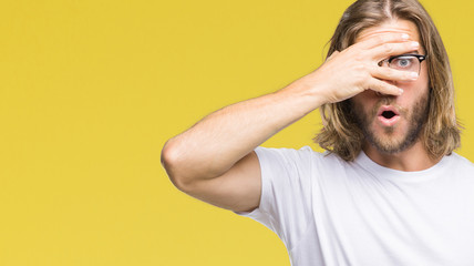 Young handsome man with long hair wearing glasses over isolated background peeking in shock covering face and eyes with hand, looking through fingers with embarrassed expression.