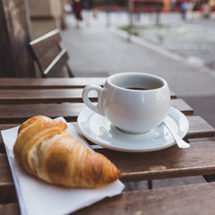 Breakfast with black coffee and croissants on the wooden table in an outdoor cafe. City on a...