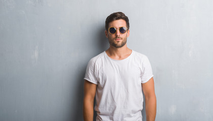 Handsome young man over grey grunge wall wearing sunglasses with serious expression on face. Simple...