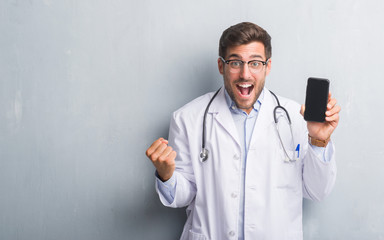 Handsome young doctor man over grey grunge wall holding smartphone screaming proud and celebrating victory and success very excited, cheering emotion