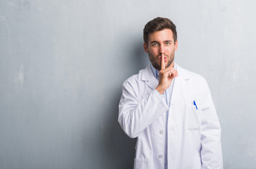 Handsome young professional man over grey grunge wall wearing white coat asking to be quiet with...