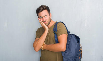Handsome tourist young man over grey grunge wall wearing backpack thinking looking tired and bored with depression problems with crossed arms.