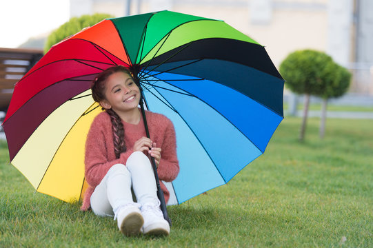Sitting under umbrella and having fun. Small girl in park waiting for rain. Colourful umbrella as concept of happiness. Rainbow that can be touched. Happy small girl smiling