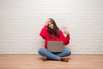 Young brunette woman sitting on the floor over white brick wall paying holding dollars stressed with hand on head, shocked with shame and surprise face, angry and frustrated. Fear and upset