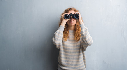 Young redhead woman over grey grunge wall looking through binoculars with a confident expression on...