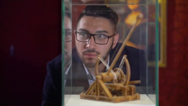 Young brunette man with a beard in glasses looks at the Da Vinci catapult model in the museum
