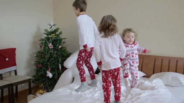 Three children dressed in Christmas pyjamas are jumping in the bed