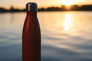Stainless red thermo bottle for water. Sunset and water on background.