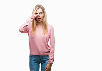 Young beautiful blonde woman wearing pink winter sweater over isolated background doing ok gesture shocked with surprised face, eye looking through fingers. Unbelieving expression.