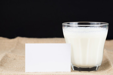 A GLASS OF MILK AND A WHITE CARD ON BROWN SACKCLOTH WITH BLACK BACKGROUND, COPY SPACE