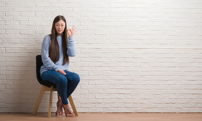 Young Chinese woman sitting on chair over brick wall doing ok sign with fingers, excellent symbol