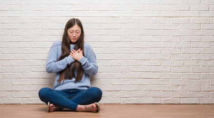 Young Chinese woman sitting on the floor over brick wall smiling with hands on chest with closed eyes and grateful gesture on face. Health concept.