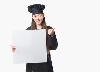 Young Chinese woman over isolated background wearing chef uniform holding banner happy with big smile doing ok sign, thumb up with fingers, excellent sign