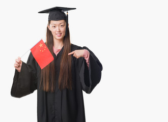 Young Chinese woman wearing graduate uniform holding China flag with surprise face pointing finger to himself