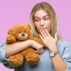 Young caucasian woman holding cute teddy bear over isolated background cover mouth with hand shocked with shame for mistake, expression of fear, scared in silence, secret concept