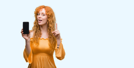 Young redhead woman showing smartphone surprised with an idea or question pointing finger with happy face, number one
