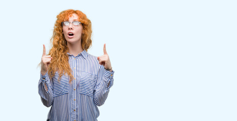 Young redhead bussines woman amazed and surprised looking up and pointing with fingers and raised arms.