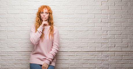 Young redhead woman standing over brick wall asking to be quiet with finger on lips. Silence and secret concept.