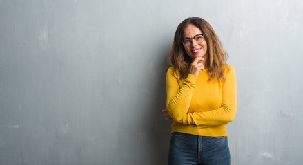 Middle age hispanic woman over grey wall wearing glasses looking confident at the camera with smile with crossed arms and hand raised on chin. Thinking positive.