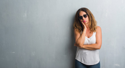 Middle age hispanic woman over grey wall wearing sunglasses thinking looking tired and bored with depression problems with crossed arms.