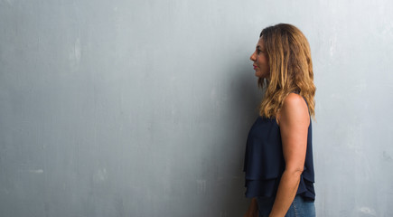 Middle age hispanic woman standing over grey grunge wall looking to side, relax profile pose with natural face with confident smile.
