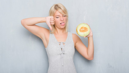 Adult caucasian woman over grunge grey wall eating cantaloupe melon with angry face, negative sign showing dislike with thumbs down, rejection concept