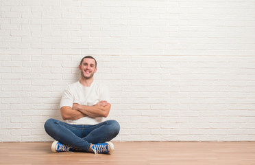 Fototapeta na wymiar Young caucasian man sitting on the floor over white brick wall happy face smiling with crossed arms looking at the camera. Positive person.