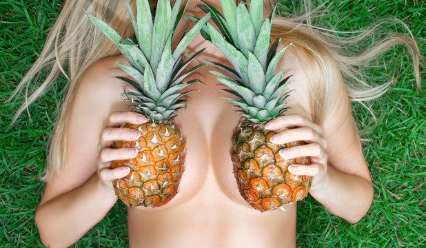 A young woman holds pineapples near her breast while lying on the grass. Bra from pineapples. Tropical modesty.