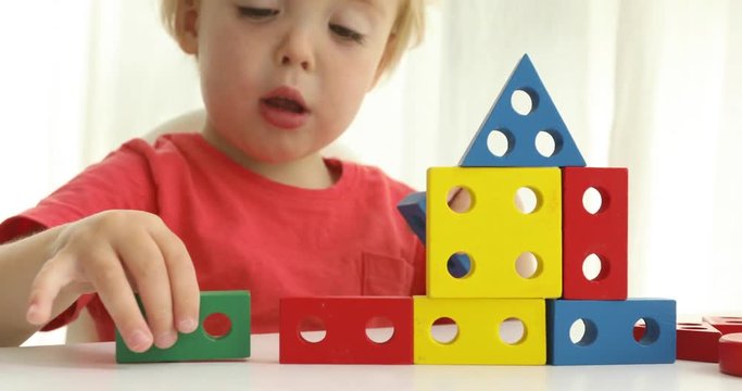 Child builds a house. Charming infant boy sitting at table and playing with colorful constructor blocks on white background
