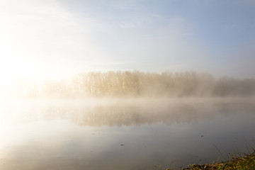 Morning autumn scenery on the bank of the river, wrapped in fog, through which the rays of the sun make their way.