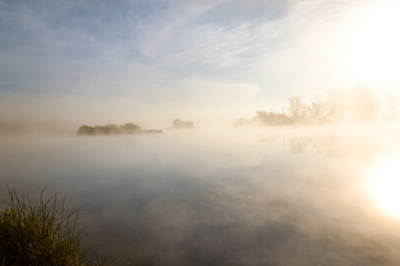 Morning autumn scenery on the bank of the river, wrapped in fog, through which the rays of the sun make their way.