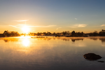 Bright orange dawn warm rays dispel mist over a calm river, illuminating the light in the area.