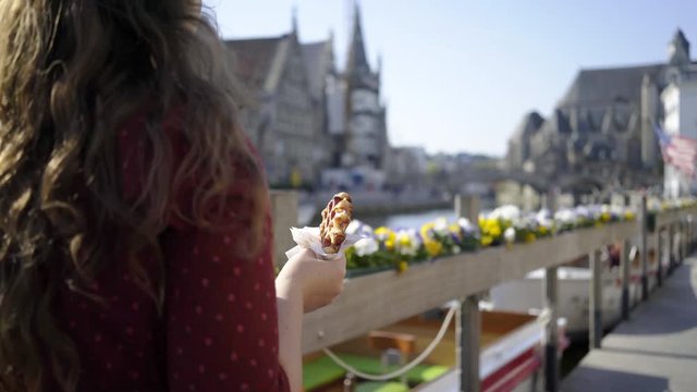 Young pretty girl walking on embankment and eat waffle. Following back view woman with belgian waffle in hand. Sunny day in Ghent, Belgium